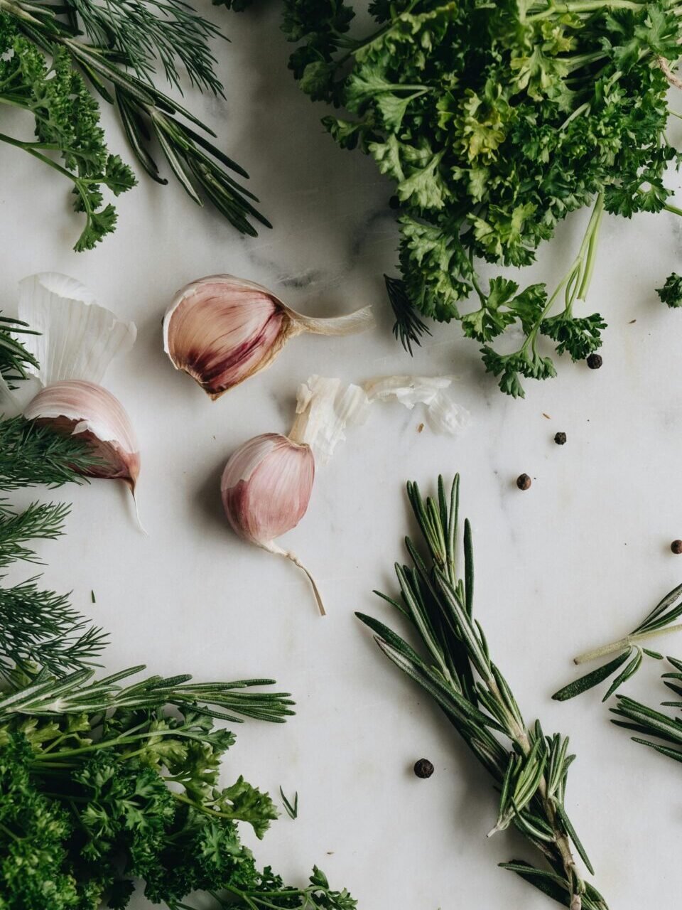 A variety of fresh herbs including garlic, parsley, and rosemary on a rustic white table, perfect for culinary use.